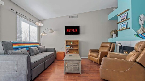 A living room with a grey sofa, a brown leather chair, a coffee table, a bookshelf, and a TV displaying 'NETFLIX'.