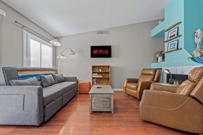 A living room with a grey sofa, a brown leather chair, a coffee table, a bookshelf, and a TV displaying 'NETFLIX'. - Dove Mountain Home | Summer Family Retreat (Tucson)