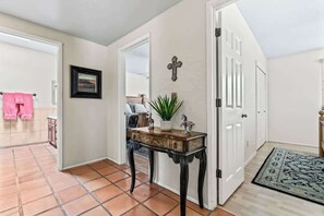 Un pasillo con suelo de baldosas, una consola de madera con una planta, una cruz en la pared y una puerta que da a otra habitación.