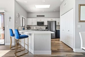 A modern kitchen with white cabinets, a central island, blue bar stools, and a stainless steel refrigerator. - Modern Foothills Summer Retreat | Mtn Views & Pool (Tucson)