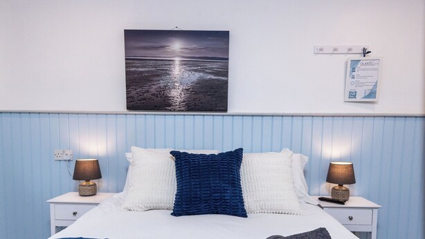 Una cama bien tendida con un edredón azul, dos lámparas de noche, un cuadro colgado en la pared y una mesita de noche blanca con una tarjeta para el huésped.