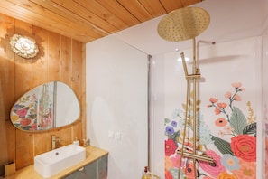 Ein Badezimmer mit Holzboden, einem weißen Waschbecken, einem großen Spiegel und einer Dusche mit floralem Dekor.