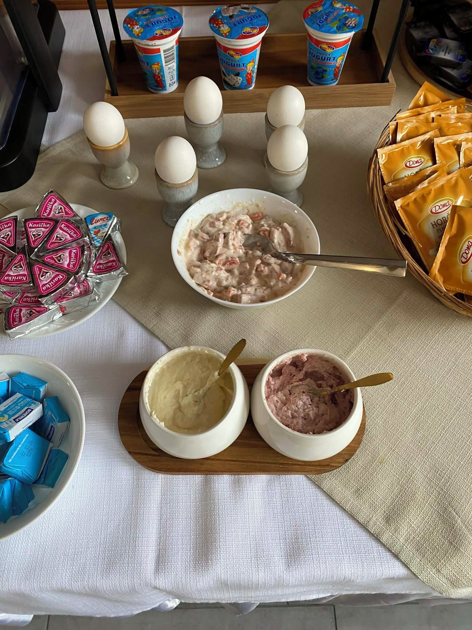 Een tafel met eten, waaronder schalen met romige en rozeachtige substanties, een bord met driehoekig verpakte snacks, en een mand met oranje zakjes.