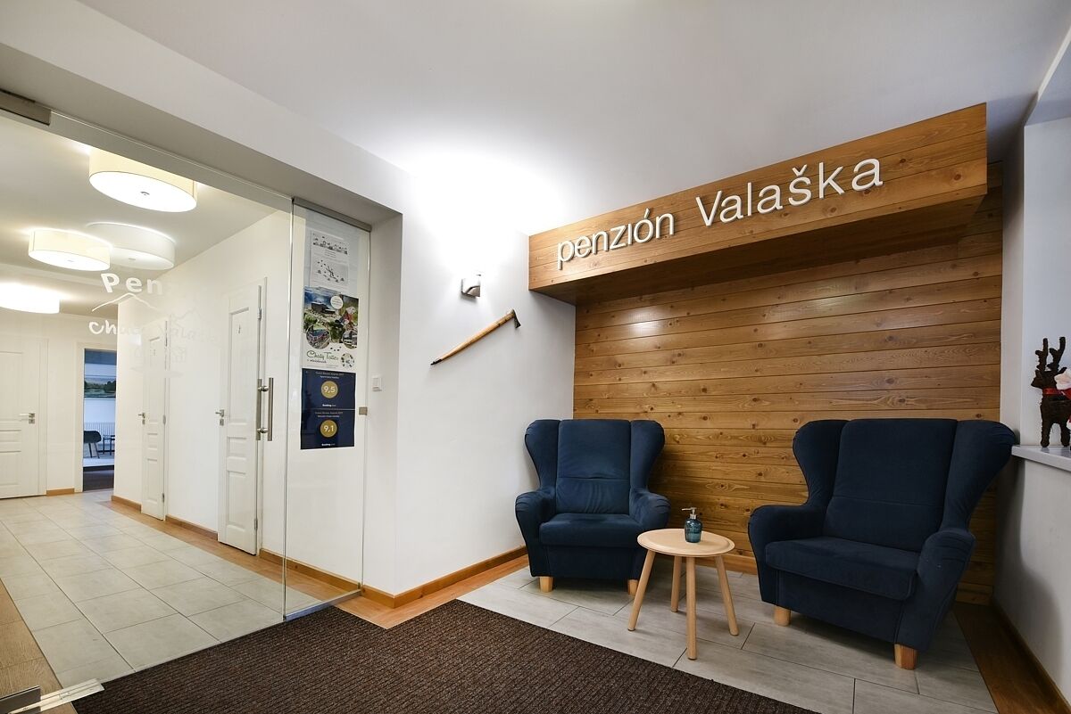 Un área de recepción con pared de madera, dos sillones azules, una pequeña mesa de madera y un letrero que dice 'pensión Valaška'.