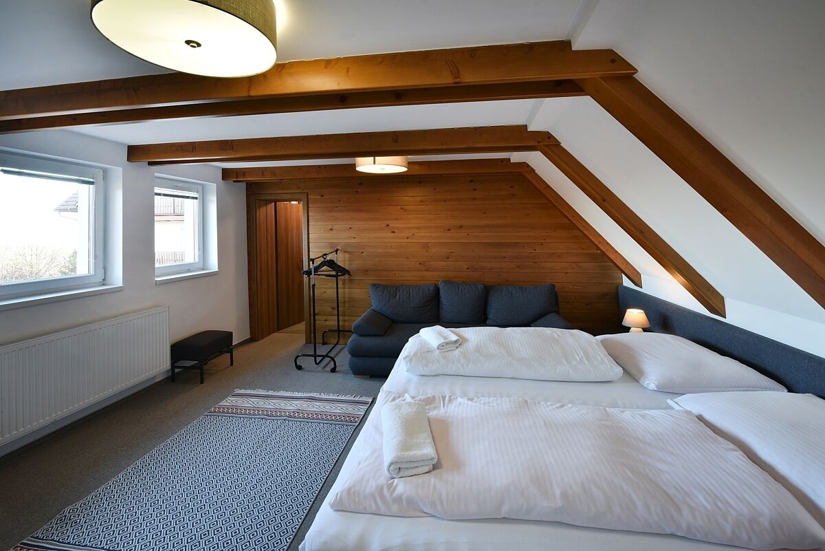 A bedroom with a sloped ceiling, a bed with white linens, a dark blue sofa, a floor lamp, and a window with a view.