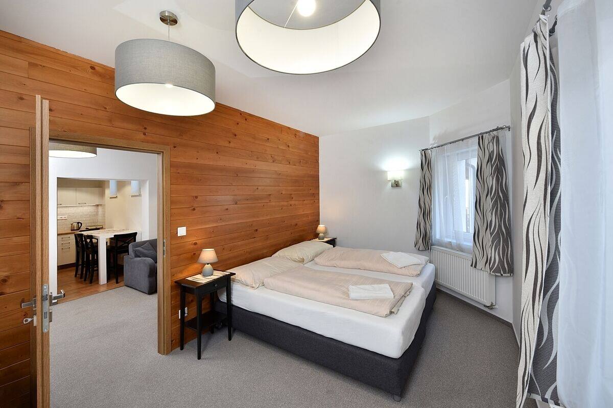 A bedroom with a wooden headboard, two beds, a nightstand, a lamp, a window with curtains, and a view into the kitchen.