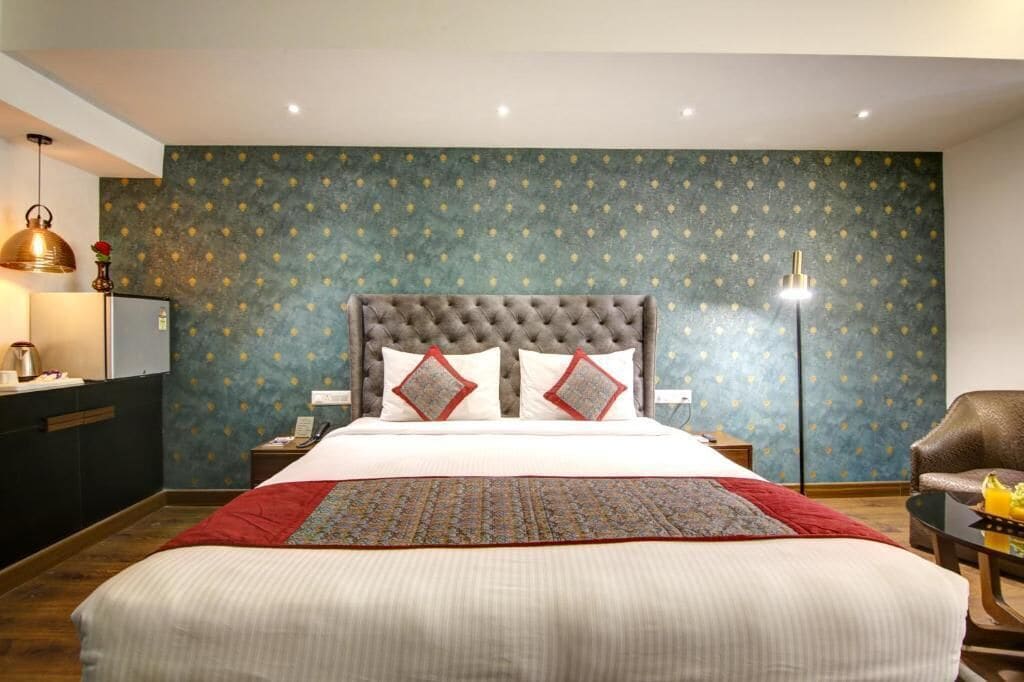 A hotel room with a large bed, a nightstand, a desk with a lamp, a chair, and a wall with a patterned wallpaper.