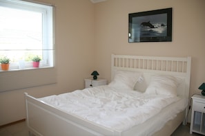 A bedroom with a white bed, a nightstand with a lamp, a window with blinds, and a framed picture on the wall. - Bungalow Near Eifel Ski Lift (Gerolstein)