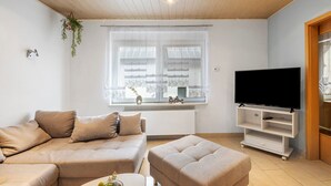A living room with a sectional sofa, a television on a stand, a window with curtains, and a hanging plant. - Holiday Home 'Family House with Huge Garden' with Sauna, Terrace and Wi-Fi (Jessen (Elster))
