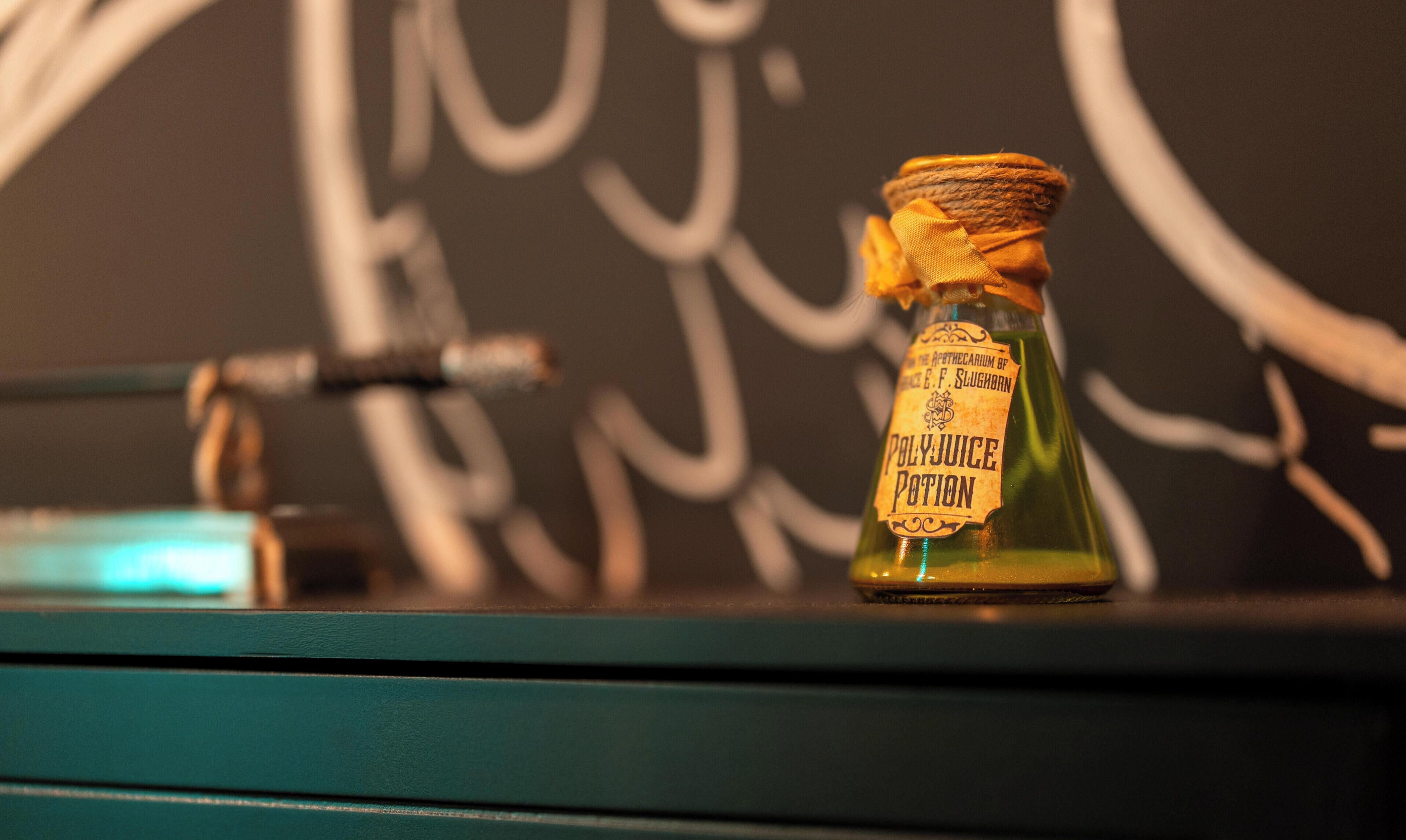 A bottle of Polyjuice Potion on a teal surface with a chalkboard in the background.