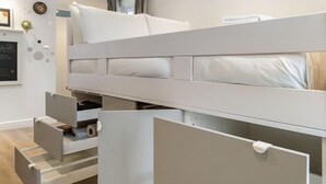 Un lit superposé avec des tiroirs de rangement en dessous, un mur blanc orné d’une horloge et d’un tableau encadré, et un plancher en bois.