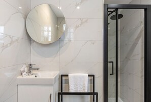 A modern bathroom with a marble wall, a round mirror, a sink, a glass shower enclosure, and a towel on a black towel rack. - GuestReady - Modern Studio in Stamford Brook (London)