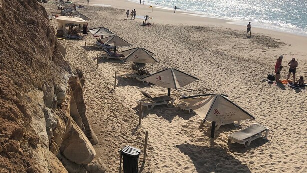 Ein Strand mit Sonnenschirmen und Liegestühlen, eine felsige Klippe und das Meer im Hintergrund.