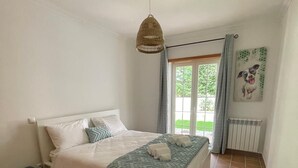Ein Schlafzimmer mit einem Bett, einem kleinen Nachttisch, einer Hängeleuchte, einem Fenster mit Vorhängen und einer Heizung.