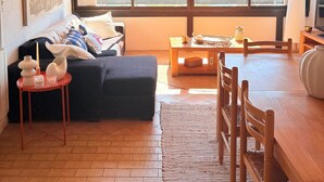 Sala de estar com sofá, mesa de centro e mesa de jantar com cadeiras. Há uma janela grande com vista para edifícios e água.