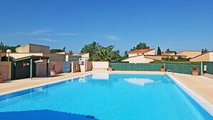 Piscina azul límpida cercada por edifícios, uma área coberta e uma cerca verde.
