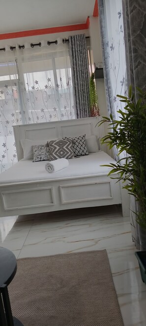 A modern bedroom with a white sofa bed, patterned pillows, a small table, and a potted plant.