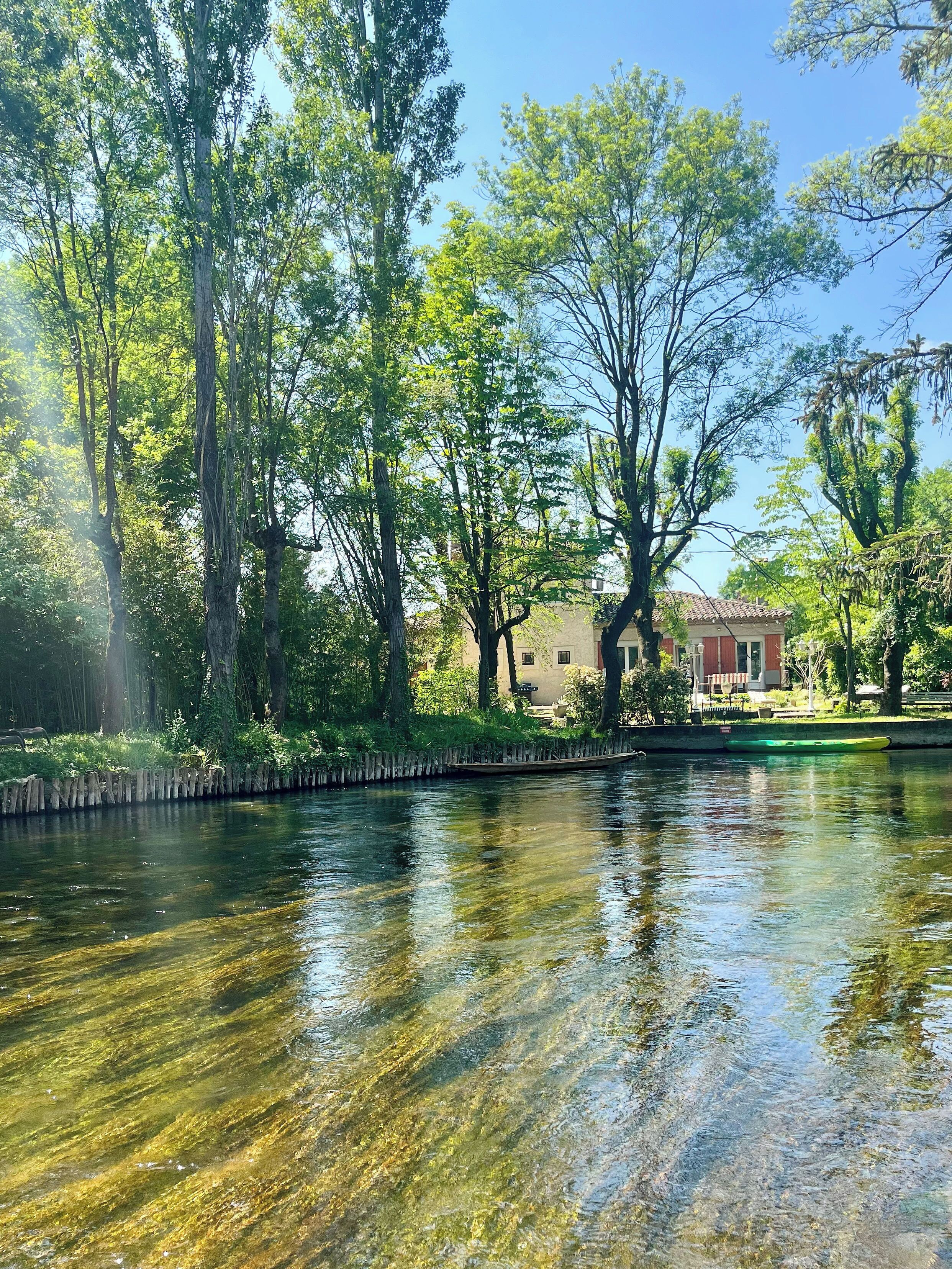 Ein ruhiger Fluss mit klarem Wasser, umgeben von üppigen grünen Bäumen und einem Gebäude mit rotem Dach im Hintergrund.