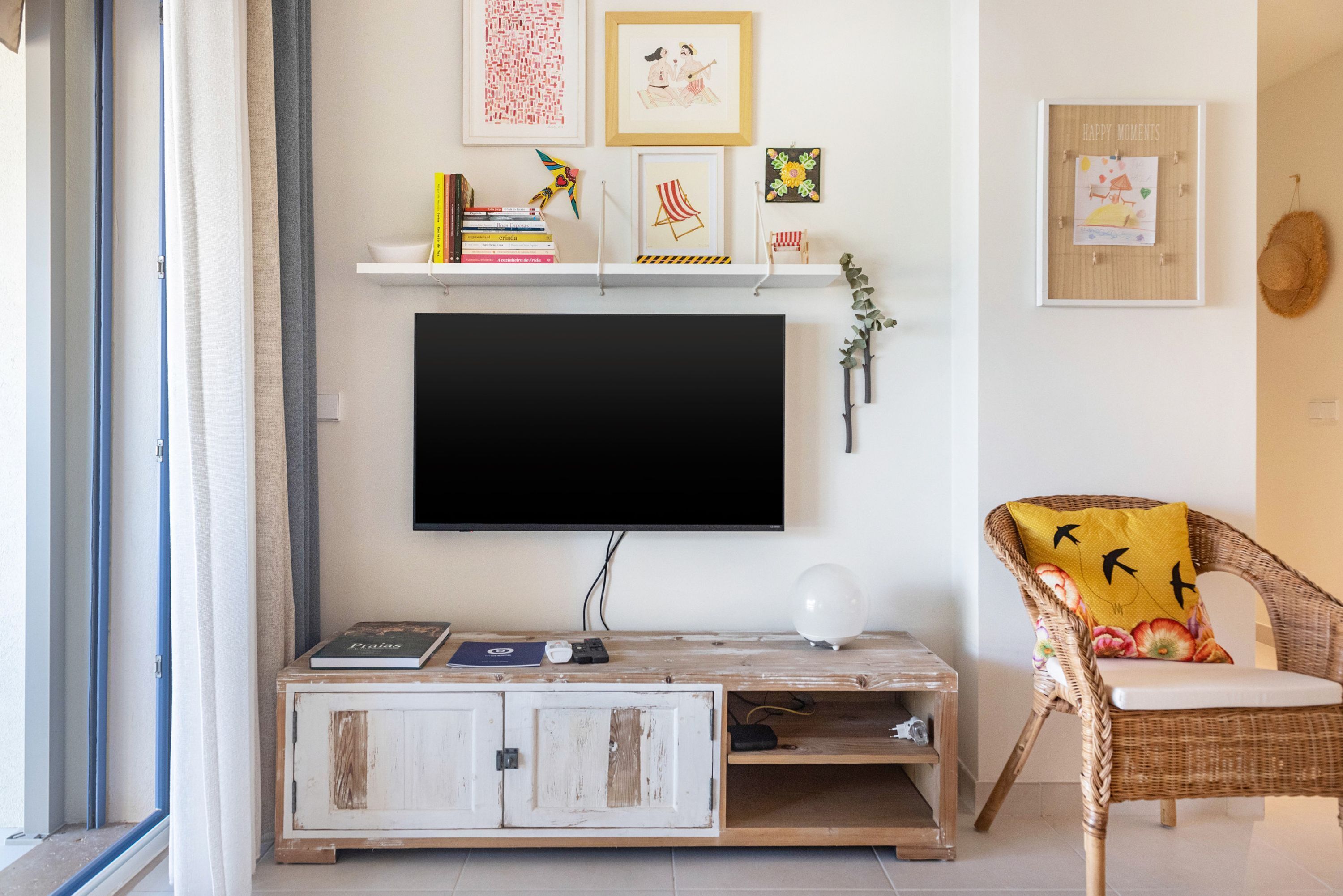 Una sala de estar con televisor de pantalla plana, una estantería con libros y objetos decorativos, un sillón de mimbre con cojín, y un marco con un cuadro colgado en la pared.