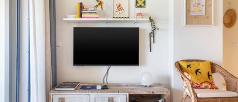 Una sala de estar con televisor de pantalla plana, una estantería con libros y objetos decorativos, un sillón de mimbre con cojín, y un marco con un cuadro colgado en la pared.
