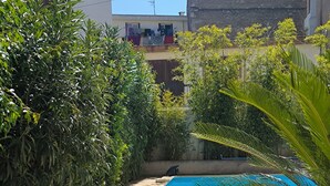 Une piscine entourée d’une végétation luxuriante, avec une terrasse en béton et un bâtiment en arrière-plan.