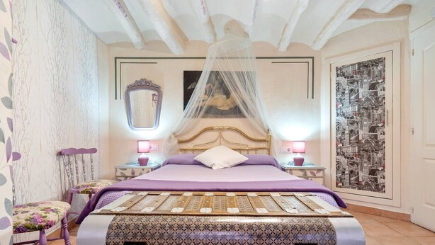Een slaapkamer met een bed, nachtkastjes, een stoel, een spiegel en een stadslandschap schilderij.