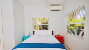 A bedroom with a bed, two bedside tables, a nightstand, a window with blinds, and an air conditioning unit. - Cozy home/Pool/Volleyball court/ near the beach (Providenciales)