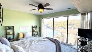 A hotel room with a bed, a ceiling fan, a TV on a stand, and a large window with a view of buildings and trees. - Stunning Lake Views at Indian Pointe! (Osage Beach)