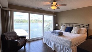 A bedroom with a bed, a chair, a ceiling fan, and a large window with a view of the water. - Beautiful Views at Bay Point Condominiums! (Osage Township)