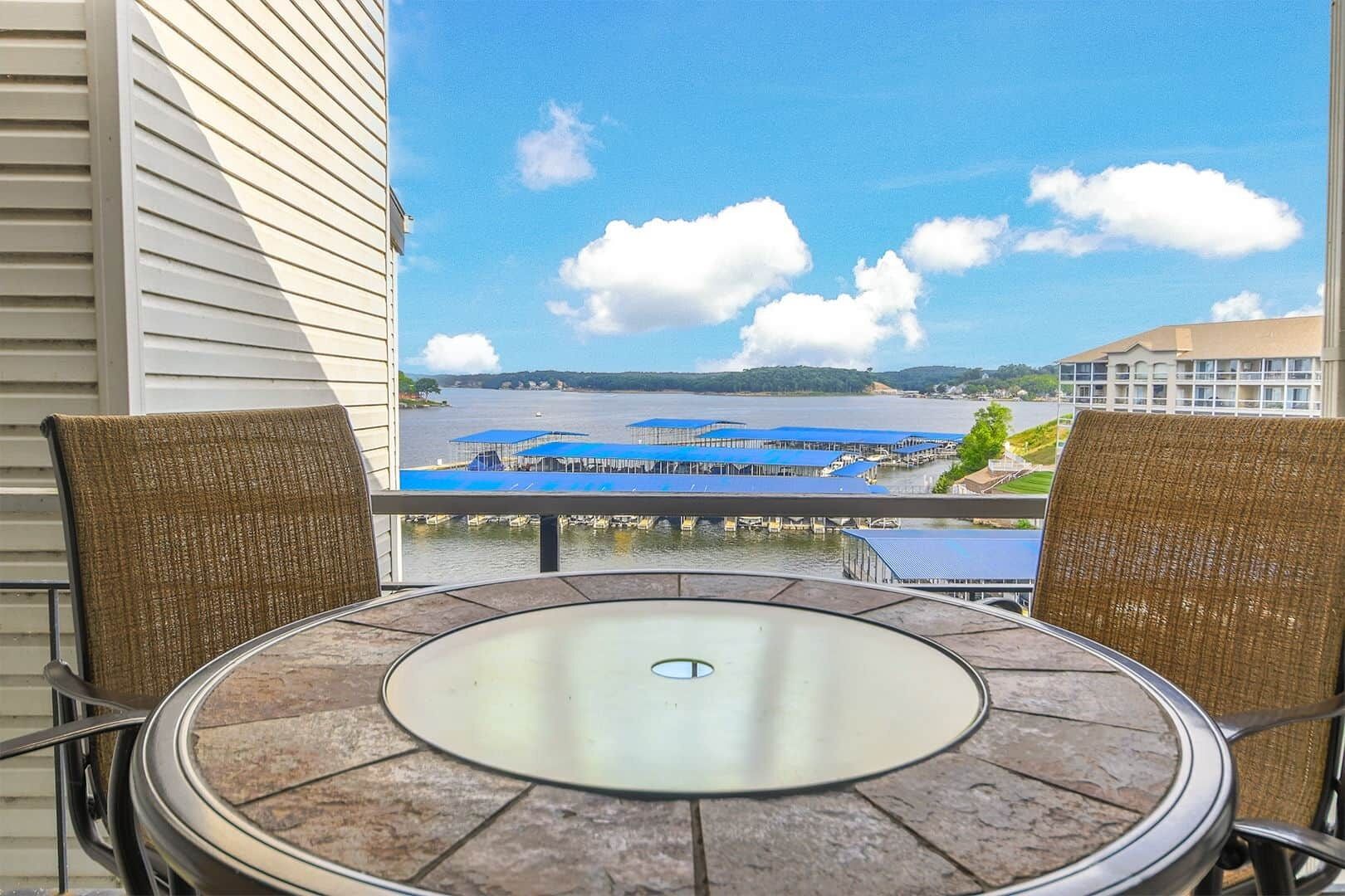 Een balkon met een ronde tafel en twee rieten stoelen, met uitzicht op een jachthaven vol boten en een verre kustlijn.