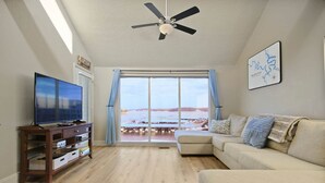 Ein Wohnzimmer mit Fernseher, einer Holz-Unterhaltungsanlage, einem beigen Sofa und einem großen Fenster mit Blick auf das Wasser.