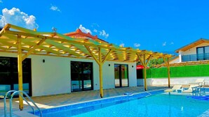 A swimming pool with a pergola, a covered patio area, and a view of a building with a green hedge. - Kent Villas Heated Pool Villas (Demre)