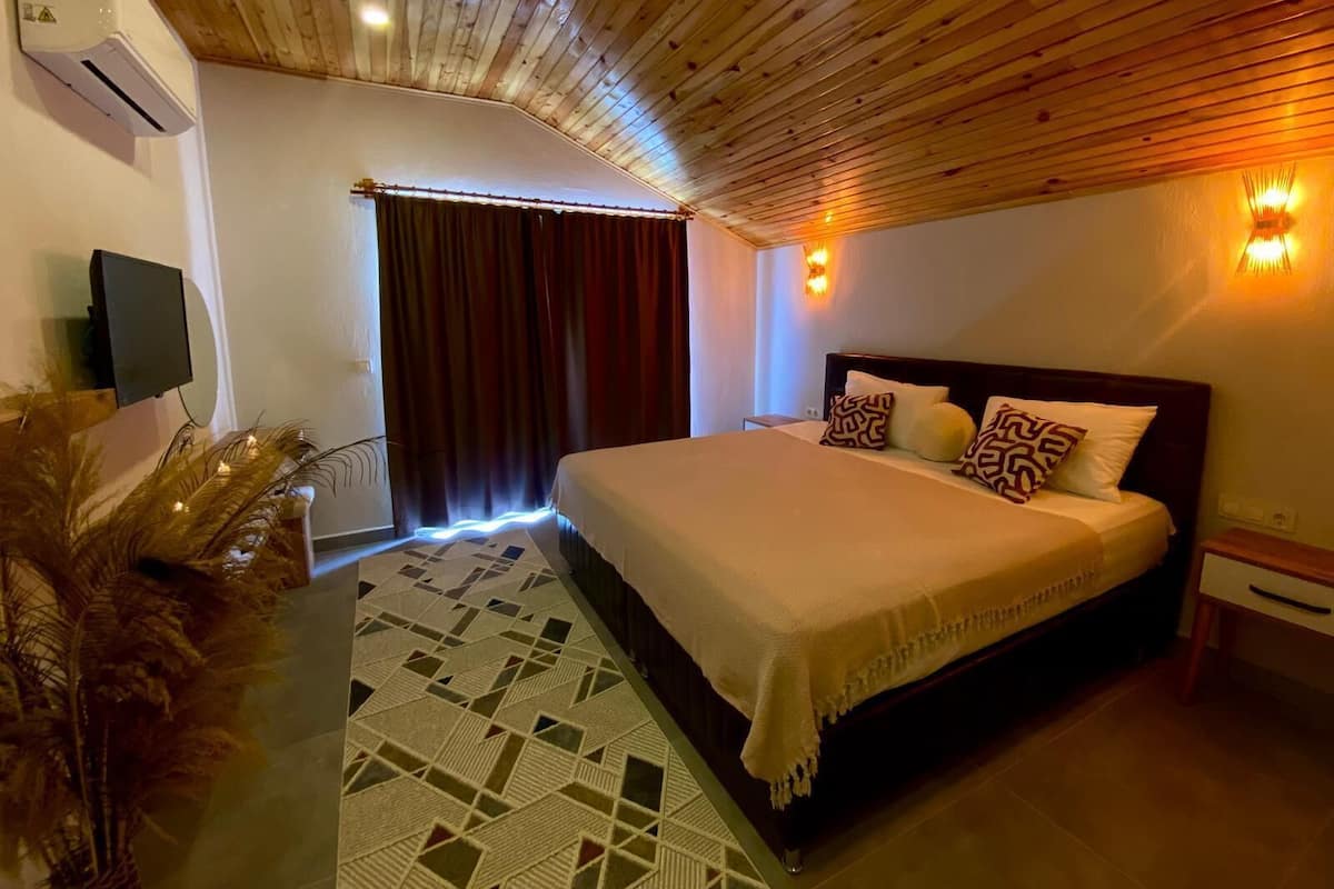 Un dormitorio con techo de madera, una cama con almohadones estampados, un televisor montado en la pared y una planta decorativa.