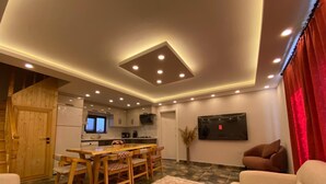 A modern living room with a wooden dining table, sofas, a TV, and a kitchen in the background. - Kent Villas Heated Pool Villas (Demre)