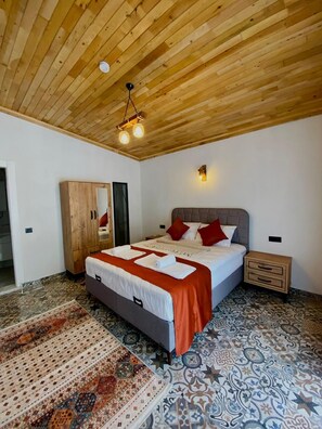 Une chambre avec un plafond en bois, un lit avec une literie blanche et des oreillers rouges, une table de chevet, une armoire et des carreaux de sol à motifs.