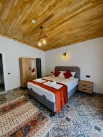 Un dormitorio con techo de madera, cama con ropa de cama blanca y almohadas rojas, mesita de noche, armario y baldosas de suelo estampadas.