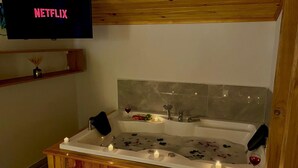 A modern bathroom with a wooden vanity, a bathtub with rose petals, candles, and a wine glass. A television displaying 'NETFLIX' is mounted above the bathtub. - Kent Villas Heated Pool Villas (Demre)