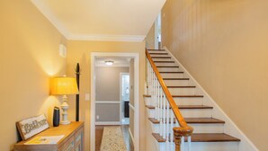 A hallway with a wooden staircase, a lamp, and a cabinet with a picture frame. - Spacious 6BR w/ Firepit, Porch, Prime Location (High Point)