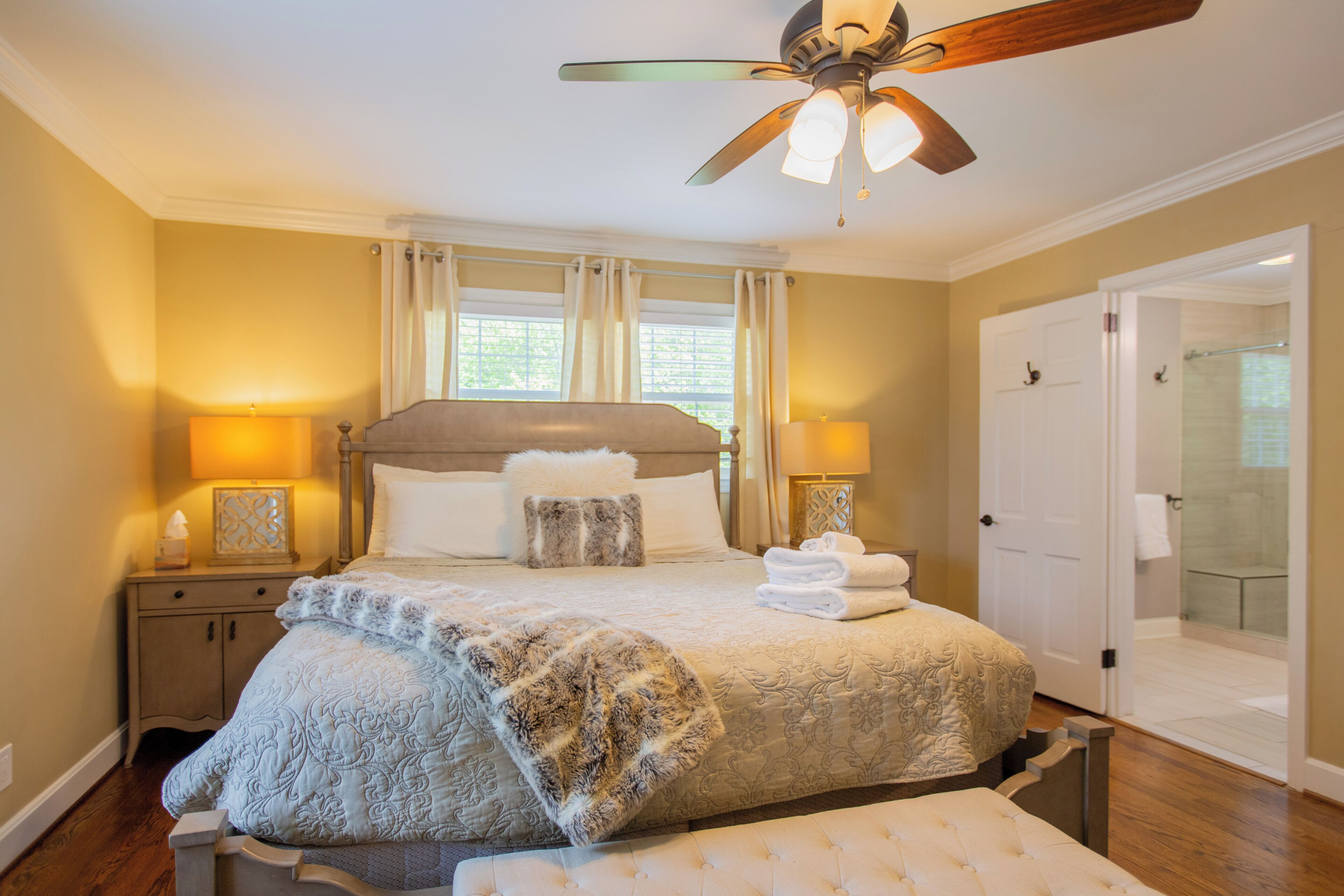 Une chambre à coucher comprenant un lit, des lampes de chevet, un ventilateur de plafond, une porte donnant sur une salle de bain et une fenêtre avec des rideaux.