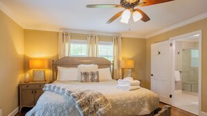 A bedroom with a bed, bedside lamps, a ceiling fan, a door to a bathroom, and a window with curtains. - Spacious 6BR w/ Firepit, Porch, Prime Location (High Point)
