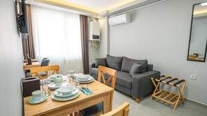 Ένα μοντέρνο σαλόνι με έναν γκρι καναπέ, ένα ξύλινο τραπέζι τραπεζαρίας στολισμένο με πιάτα και ποτήρια, ένα μικρό ξύλινο βοηθητικό τραπεζάκι και μια μονάδα κλιματισμού τοποθετημένη στον τοίχο.