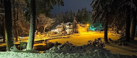 A snow-covered landscape with a building illuminated by warm lights, surrounded by trees.