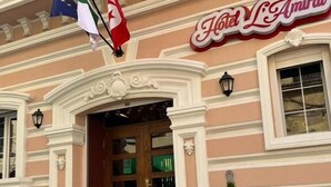 A building with a sign reading 'Hotel L'Amiral,' a wooden door, and two potted plants. Flags are visible above the entrance. - L amiral (Oran)
