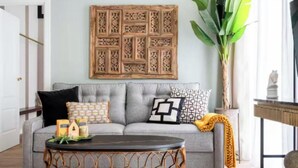 Ein Wohnzimmer mit grauem Sofa, gemusterten Kissen, einem Holzbild, einem runden Tisch und einer großen Pflanze.