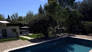 A rectangular swimming pool with a wooden deck surrounding it, set in a landscaped area with trees and a small building in the background. - Mazet Léa - Quiet terrace & pool (Le Rouret)