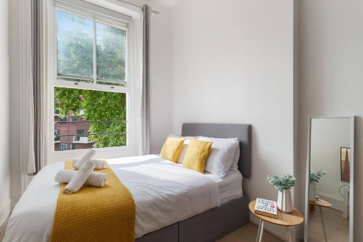 A hotel room with a bed, a window with a view of greenery, a bedside table with a book, and a mirror.