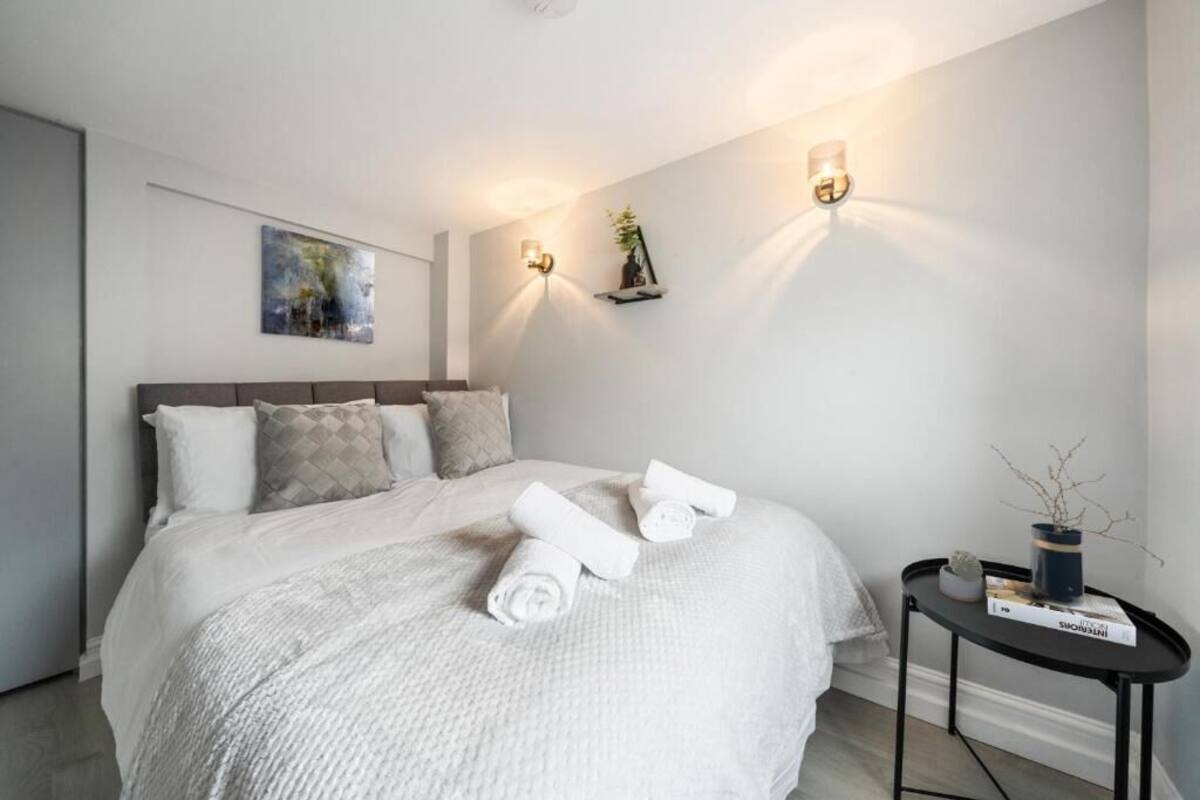 A modern bedroom with a large bed, two wall-mounted lights, a small side table with a plant, and a painting on the wall.