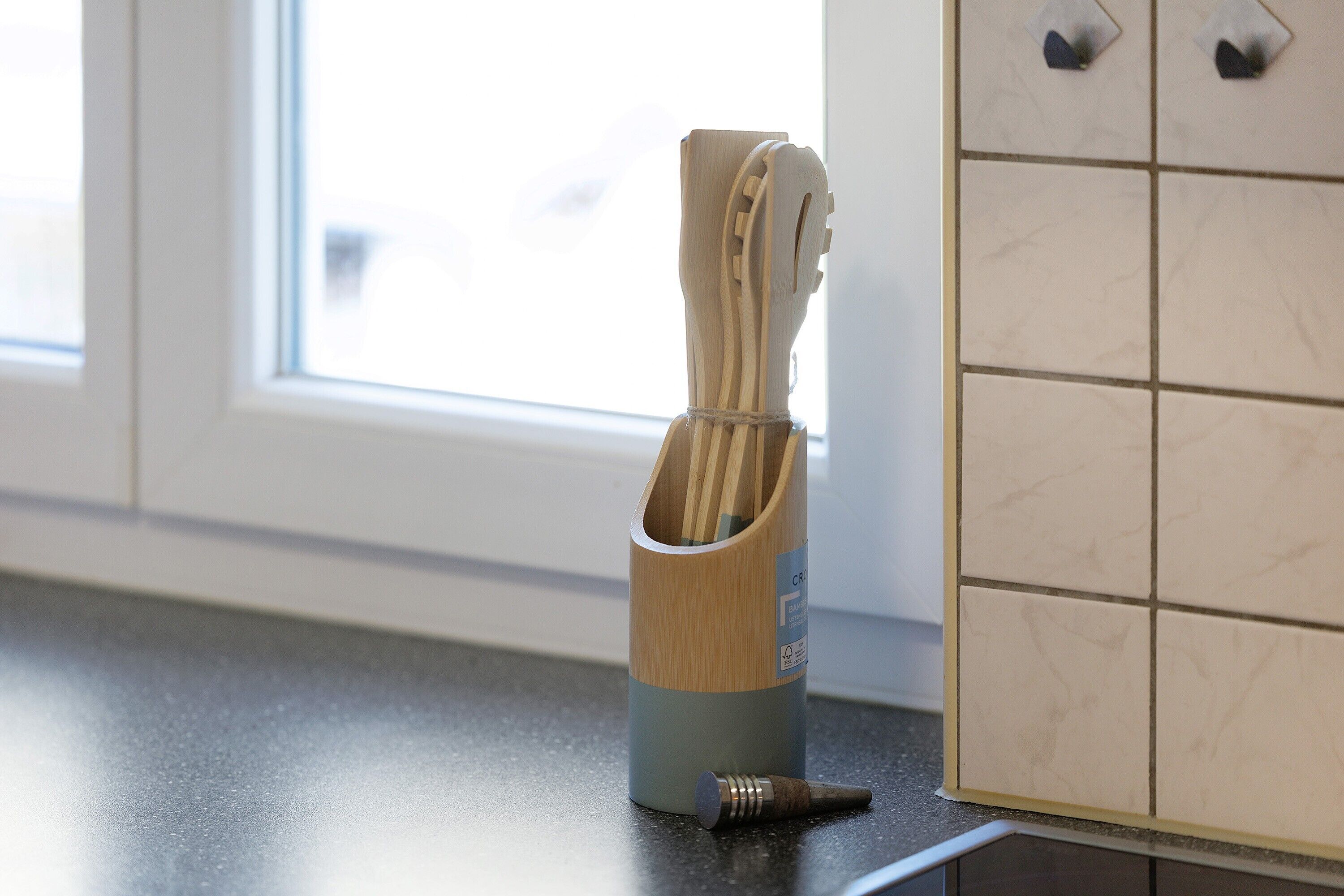 Eine Küchenarbeitsplatte mit einem Utensilienhalter, der Holzkocheräe enthält, ein Fenster und eine geflieste Spritzschutzwand.