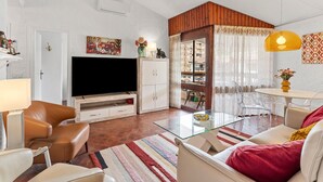 Una sala de estar con televisor, sillones de cuero, una mesa de centro de vidrio y un comedor con una lámpara colgante amarilla.