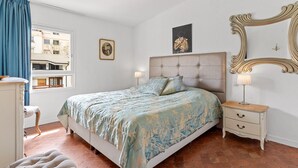 Un dormitorio con cama, mesita de noche, espejo y ventana con cortinas.